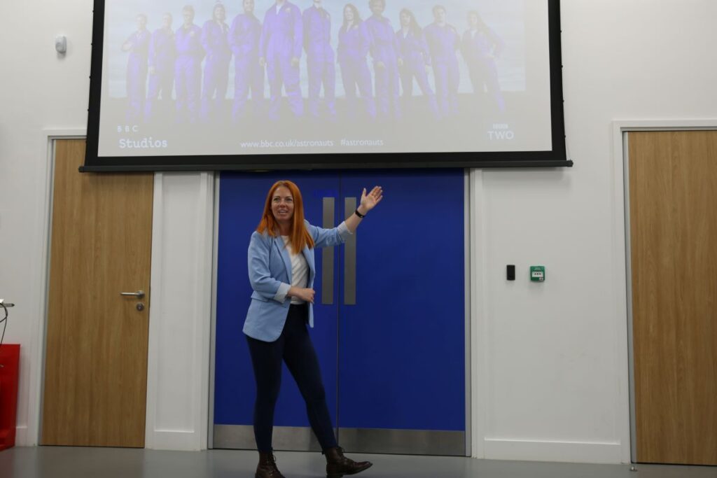 Photo of Dr Jackie Bell in gesturing in front of screen with photo of her and group of people