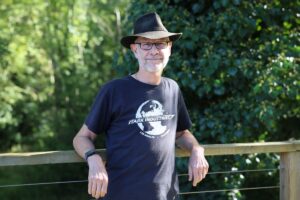 Head and upper body shot of Graham Quelch, wearing hat, glasses and black t-shirt posed in front of greenery