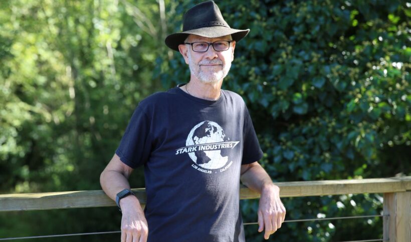 Head and upper body shot of Graham Quelch, wearing hat, glasses and black t-shirt posed in front of greenery
