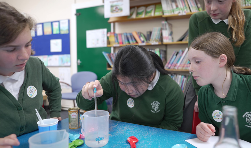 Pupil measuring temperature of liquid in a plastic beaker with a thermometer, with three other pupils looking on.