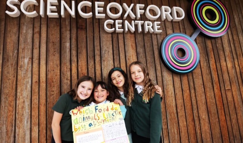 Group of 4 children holding cardboard poster in front of building with signage of Science Oxford Centre