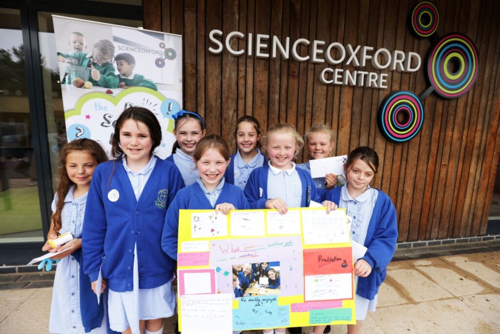 group of 7 children holding cardboard poster in front of building with signage of Science Oxford Centre
