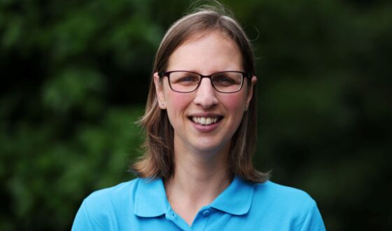 Head shot of woman with dark blonde medium length hair, wearing glasses, against green background out of focus