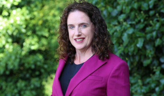 head and shoulder photo of Caucasian woman with long brown curly hair, wearing purple blazer over black top.