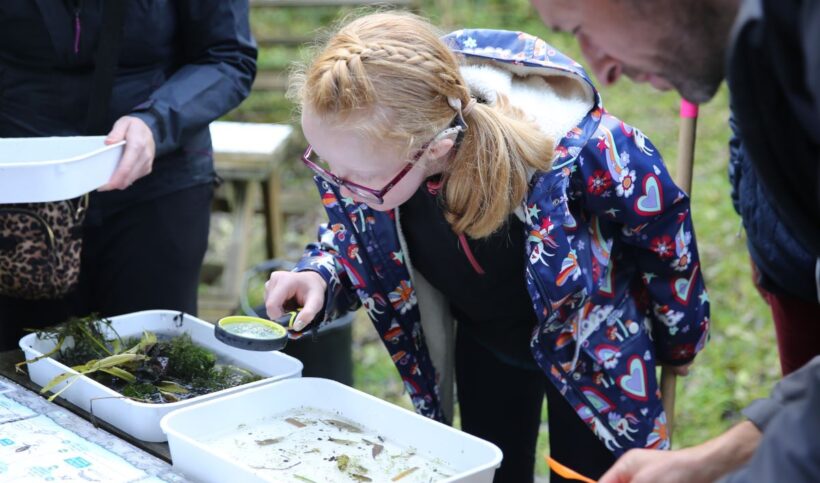 Girl holding a magnifying glass, bending over to look into a container with water and insects.