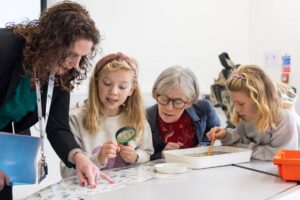 Two adults and two children looking at pond water in white flat tub and pointing at insects on chart